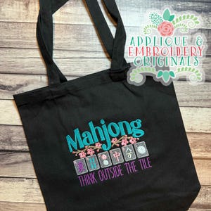 K&ouml;nnte beinhalten: Schwarze Tragetasche mit t&uuml;rkisfarbenem gesticktem Schriftzug "Mahjong", rosa Kirschbl&uuml;ten und Mahjong-Steinen. Der Satz "THINK OUTSIDE THE TILE" ist lila gestickt. Die Tasche hat schwarze Griffe und ist aus robustem Stoff gefertigt.