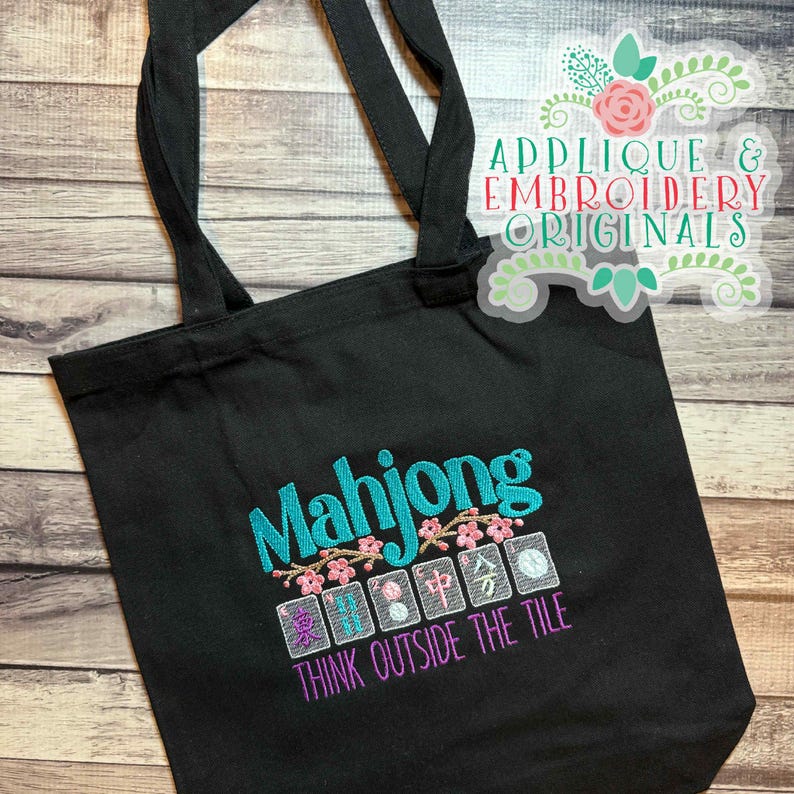 K&ouml;nnte beinhalten: Schwarze Tragetasche mit t&uuml;rkisfarbenem gesticktem Schriftzug "Mahjong", rosa Kirschbl&uuml;ten und Mahjong-Steinen. Der Satz "THINK OUTSIDE THE TILE" ist lila gestickt. Die Tasche hat schwarze Griffe und ist aus robustem Stoff gefertigt.