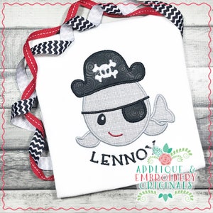 Puede incluir: Una camiseta blanca con una ballena gris que lleva un sombrero de pirata negro con una calavera y huesos cruzados. La ballena tiene un parche en el ojo negro y una sonrisa roja. El nombre "LENNO" está bordado debajo de la ballena.