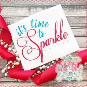 Puede incluir: Tela blanca con bordado rojo y azul que dice "it's time to sparkle". La tela está rodeada de una cinta roja y cuentas de perlas blancas. La imagen también incluye un logotipo para Applique & Embroidery Originals.