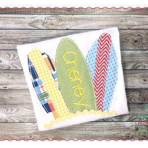 May include: A white t-shirt with three colorful surfboards appliqued on the front. The middle surfboard has the name "Cheney" embroidered on it in yellow thread.