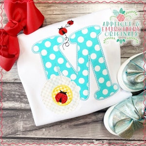 Op de afbeelding: Een witte romper met een turquoise letter M met witte stippen. Een witte bloem met een gele kern en een rood lieveheersbeestje bevindt zich onder de letter. Drie rode lieveheersbeestjes bevinden zich boven de letter. De tekst "Applique & Embroidery Originals" bevindt zich in de rechterbovenhoek van de afbeelding.