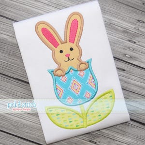 Puede incluir: Diseño bordado de un conejito beige con orejas rosas que emerge de un tulipán azul estampado. El tulipán tiene hojas verdes y amarillas, sobre un fondo blanco. El texto "pick & stitch" está en la parte inferior.