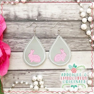 May include: A pair of gray teardrop-shaped earrings with pink bunny rabbit embroidery. The earrings have silver hooks.