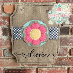 May include: A burlap welcome sign with a pink flower applique, black and white checkered fabric, and the text "welcome". The sign is decorated with a blue and white flower with a yellow center. The sign also includes the text "Applique & Embroidery Originals".