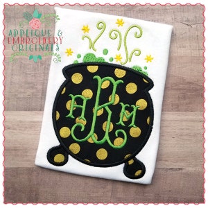 May include: A black cauldron with gold polka dots and green initials "AKM" is embroidered on a white shirt. Green swirls and yellow stars are embroidered above the cauldron.