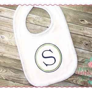May include: A white baby bib with a white trim and a green and blue embroidered circle with the letter "S" in the centre. The bib is on a wooden background. Applique Embroidery Originals.