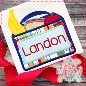 Könnte beinhalten: Eine blaue Lunchbox mit einem roten Apfel, einem Sandwich und einer gelben Banane. Die Lunchbox hat ein kariertes Muster und ein blaues Namensschild mit der Aufschrift "Landon".