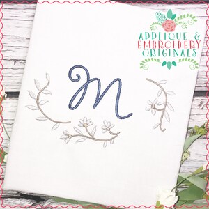 Könnte beinhalten: Stickerei mit dem Buchstaben "M" in Blau und einem floralen Kranz in Braun- und Weißtönen. Das Design ist auf einem weißen Stoffhintergrund. Der Text "Applique & Embroidery Originals" befindet sich in der oberen rechten Ecke.