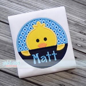 Puede incluir: Un artículo de tela blanca con un diseño bordado de un pollito amarillo en un círculo con estampado azul y blanco. El pollito tiene cuerpo amarillo, ojos negros y pico naranja. El nombre "Matt" está bordado en azul claro debajo.