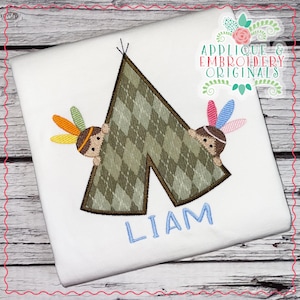 May include: White t-shirt with an embroidered teepee design. The teepee is green with a brown argyle pattern and two cartoon Native American figures peeking out. The name "LIAM" is embroidered in blue below the teepee. The text "APPLIQUE & EMBROIDERY ORIGINALS" is in the upper right corner.