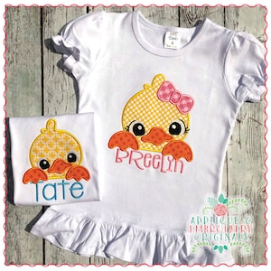 May include: Two white t-shirts with embroidered yellow duck faces. The duck on the larger shirt has a pink bow. The smaller shirt has the name "Tate" embroidered on it. The larger shirt has the name "Preelin" embroidered on it.