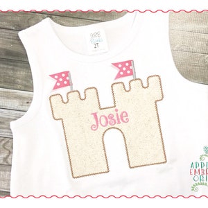 May include: A white sleeveless shirt with a beige sandcastle applique with pink flags and the name "Josie" embroidered on it.