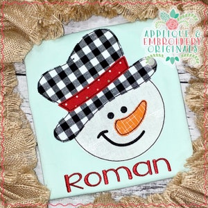 May include: A white snowman with a black and white checkered hat, red and white trim, and a carrot nose. The snowman is smiling and the name "Roman" is embroidered below it.