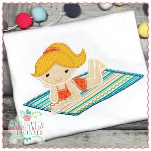 May include: Embroidered design of a girl with blonde hair wearing a swimsuit and lying on a striped beach towel.