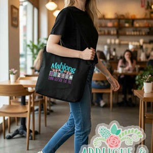 K&ouml;nnte beinhalten: Schwarze Tragetasche mit dem Wort "Mahjong" in T&uuml;rkis und Lila, mit dem Satz "Think Outside the Tile". Die Tasche wird von einer Person getragen, die ein schwarzes T-Shirt und blaue Jeans tr&auml;gt.