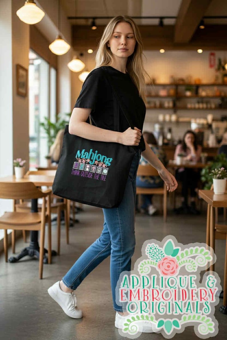 K&ouml;nnte beinhalten: Schwarze Tragetasche mit dem Wort "Mahjong" in T&uuml;rkis und Lila, mit dem Satz "Think Outside the Tile". Die Tasche wird von einer Person getragen, die ein schwarzes T-Shirt und blaue Jeans tr&auml;gt.