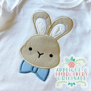 Puede incluir: Body blanco con un aplique de conejo beige y una pajarita azul con lunares blancos. El conejo tiene ojos negros y orejas beige con el interior blanco. El aplique está bordado con hilo beige claro. El texto "APPLIQUE & EMBROIDERY ORIGINALS" está en la esquina inferior derecha.