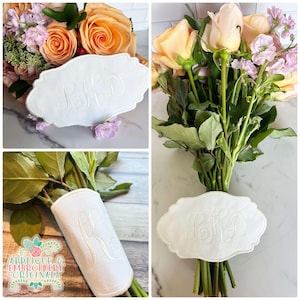 May include: White fabric bouquet wrap with a monogrammed design. The wrap is tied around a bouquet of peach and pink roses and greenery.
