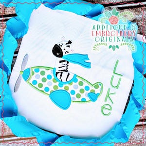 May include: White embroidered applique of a zebra wearing a blue scarf and flying a blue and green airplane with white polka dots. The name "Luke" is embroidered in green below the airplane.
