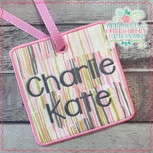May include: Pink and white striped fabric tag with the names "Charlie Kate" embroidered in gray thread. The tag has a pink border and a hole at the top for hanging.