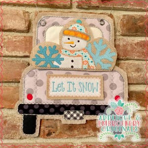May include: A gray and white truck with a snowman holding a snowflake in the back. The truck has a sign that says "Let It Snow".