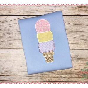 Peut inclure: Un t-shirt bleu clair avec une application de cornet de glace. Le cornet de glace a trois boules roses, jaunes et violettes, avec une base en gaufre. L'application est cousue sur le t-shirt.
