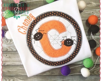 Könnte beinhalten: Ein weißes T-Shirt mit einem schwarzen und orangen Kreisdesign, das ein Spinnennetz und zwei Spinnen zeigt. Der Buchstabe "C" befindet sich in der Mitte des Kreises und der Name "Chevy" ist über dem Kreis gestickt.