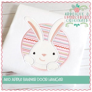 Puede incluir: Colgador de puerta de tela blanca con un diseño de conejo bordado. El conejo está dentro de un círculo rosa y a rayas. El texto "Aeo APPLE BANNER DOOR HANGER" está en la parte inferior. El logotipo "Applique & Embroidery Originals" está en la parte superior.