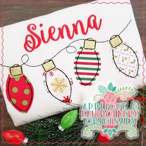 May include: A white fabric with the name "Sienna" embroidered in red cursive lettering. A string of five Christmas lights is embroidered below the name. Each light is a different pattern, including red and green polka dots, a snowflake, red and green stripes, and a red and green plaid.