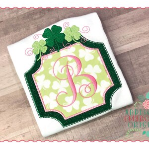 May include: A white shirt with a green shamrock design and a pink letter "B" in a green frame. The design is surrounded by green shamrocks and swirls.