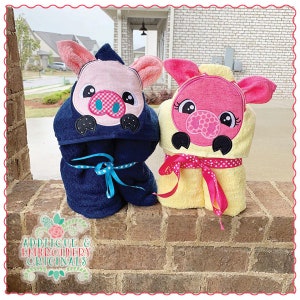 May include: Two hooded towels with pig faces embroidered on them. The towels are blue and yellow with pink pig faces. The pig faces have black eyes, pink noses, and white eyelashes.