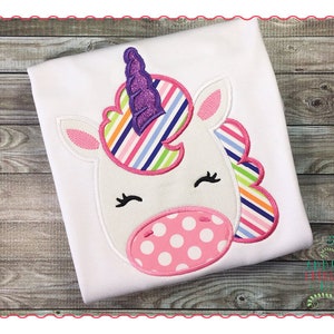 May include: White unicorn applique with a pink and white polka dot nose, rainbow striped mane, and a purple sparkly horn. The applique is sewn onto a white t-shirt.