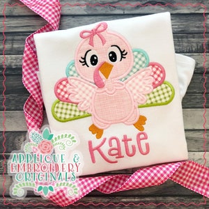 May include: A pink and white applique of a turkey with a bow on its head. The turkey is embroidered on a white shirt with the name "Kate" embroidered below it.