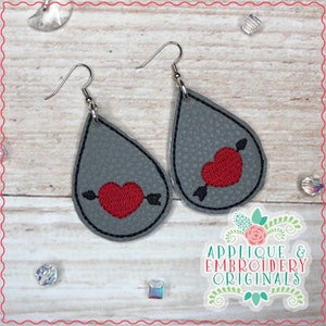 May include: A pair of gray teardrop earrings with a red heart pierced by a black arrow. The earrings have a black outline and silver hooks. The image also includes the text "Applique & Embroidery Originals".