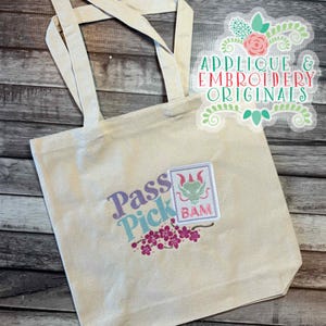 Puede incluir: Un bolso tote de lona beige con las palabras "Pass Pick" en escritura morada y turquesa. Un parche con un diseño de dragón verde y la palabra "BAM" está en la parte delantera, junto con bordados florales rosas. El bolso tiene dos asas beige.
