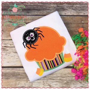 May include: A white t-shirt with an embroidered orange cupcake with a black spider on top. The cupcake has a striped icing and candy corn on the side.