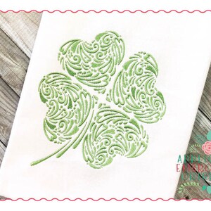 May include: A green four-leaf clover with a swirling, intricate design. The clover is embroidered on a white fabric background.