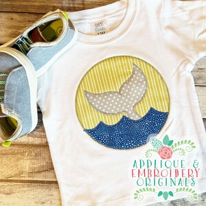 May include: White short-sleeved t-shirt with a whale tail applique design. The design features a gray whale tail with white polka dots, blue waves, and a yellow striped background. A pair of blue and gray shoes are next to the shirt. The shirt has the text "Applique & Embroidery Originals".