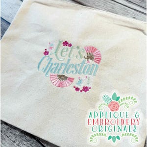 Op de afbeelding: Crèmekleurige draagtas met geborduurde tekst "Let's Charleston" in lichtblauw en groen, omringd door roze waaier- en bloemmotieven. Een sticker met de tekst "Applique & Embroidery Originals" staat in de rechterbenedenhoek.