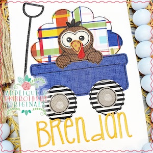 May include: A blue wagon with a turkey inside. The turkey is brown with a red wattle and a plaid pattern on its back. The wagon has black and white striped wheels and the name "Brendan" is embroidered below.