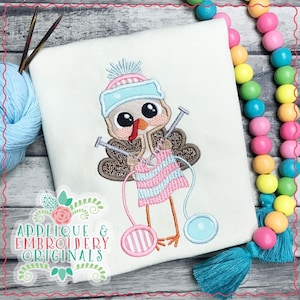 Puede incluir: Diseño bordado de un pavo que lleva un gorro y una bufanda azules y rosas, sosteniendo agujas de tejer y lana. El pavo está tejiendo una bufanda de rayas rosas y azules.
