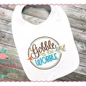 May include: White baby bib with a brown circle and the text "Gobble 'til you Wobble" in colorful script. A colorful arrow is inside the circle.