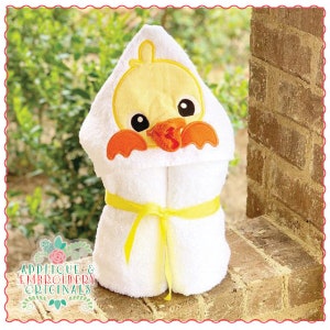 Könnte beinhalten: Ein weißes Kapuzenhandtuch mit einer gelben Entenapplikation. Die Ente hat einen orangefarbenen Schnabel und Füße. Das Handtuch hat ein gelbes Band darum gebunden.