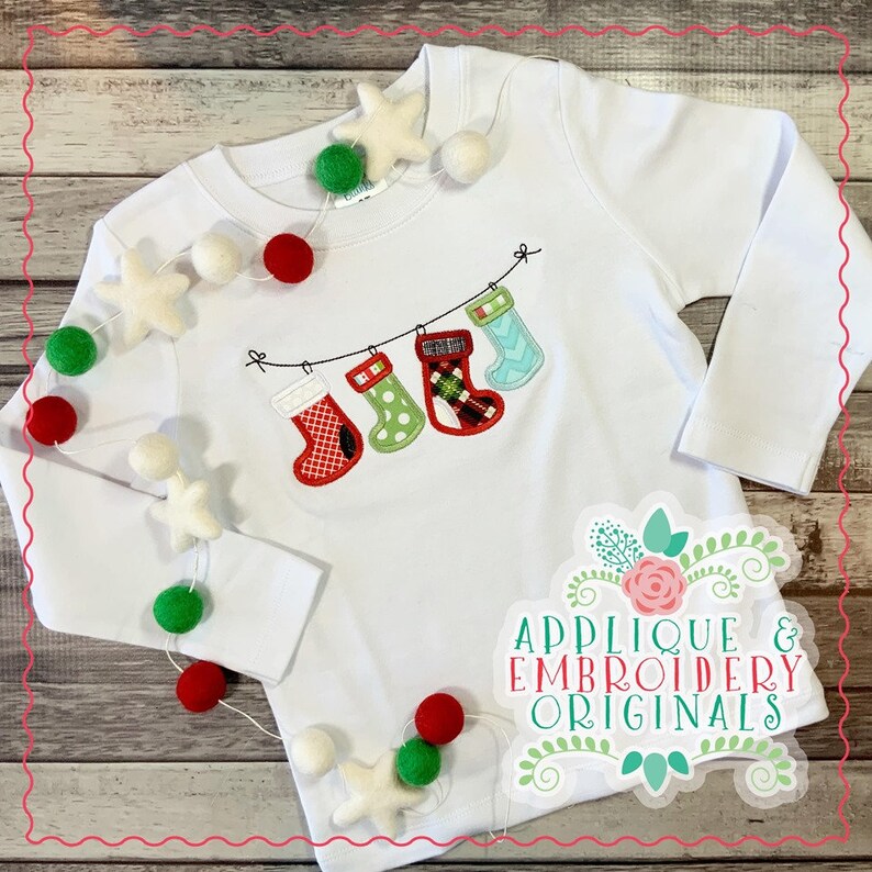 May include: White long-sleeved shirt with an embroidered design of Christmas stockings hanging on a string. The shirt is decorated with a garland of white and red pom-poms and white felt stars.