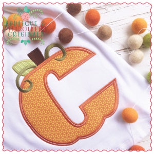 May include: A white fabric with an orange pumpkin-shaped letter C. The letter is outlined in brown and has a green stem and leaf. The fabric is surrounded by small, colorful pom-poms.