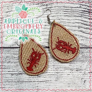 May include: A pair of teardrop-shaped earrings with a red embroidered lobster design on a burlap background. The earrings are made of metal and have a hook fastening.
