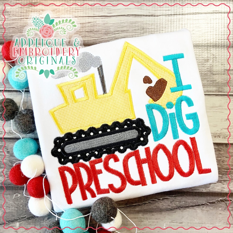 May include: A white t-shirt with an embroidered yellow excavator, a brown heart, and the text "I Dig Preschool" in red, blue, and turquoise.