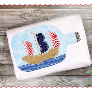 Peut inclure: Un t-shirt blanc avec une applique bleue, rouge et blanche d'un voilier dans une bouteille. Le voilier a une voile rayée rouge, blanche et bleue, une voile bleue à pois blancs et une voile grise à pois blancs.