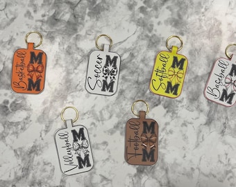 May include: Keychains with gold-colored rings and rectangular tags. Each keychain features a different sport: basketball, soccer, softball, baseball, volleyball, and football. The word "MOM" is displayed on each keychain, along with a sport-related graphic.